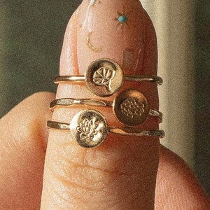 以下が含まれることがあります： 指に重ねられた、円形のエンボス加工が施された4つのゴールドカラーのリング。デザインには、蓮の花、複数の花びらを持つ花、および他の2つの抽象的なデザインが含まれています。リングは薄く繊細で、研磨された仕上げです。