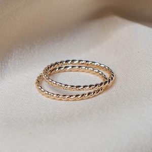 May include: Two gold-colored, twisted rope design rings. The rings are circular and sit on a cream-colored fabric. The rings are made of a shiny, metallic material. The rings are likely jewelry.