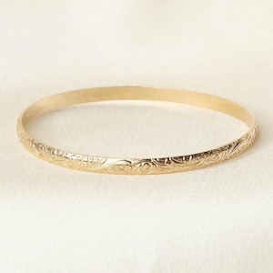 May include: A gold-colored bangle bracelet with an engraved floral pattern. The bracelet is circular and appears to be made of metal. The floral design is repeated around the entire circumference of the bracelet.