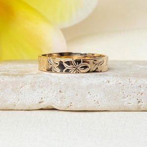 Può includere: Un anello color oro con un motivo floreale è appoggiato su una pietra di colore chiaro. Il design dell'anello presenta motivi floreali ripetuti attorno alla sua fascia. Lo sfondo include un fiore giallo tenue e una superficie bianca, creando un'estetica delicata ed elegante.