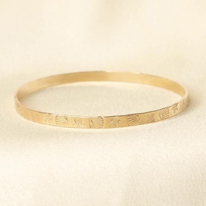 May include: A gold-coloured bangle bracelet featuring a repeating pattern of seashells and starfish. The thin, circular bracelet has the design etched into the metal. The background is a soft, off-white fabric.