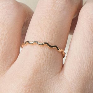 May include: A gold-colored ring with a wavy design. The ring is thin and delicate, featuring a continuous wave pattern around the band. The ring is shown on a finger, highlighting its simple yet elegant style. This ring is a piece of jewelry.