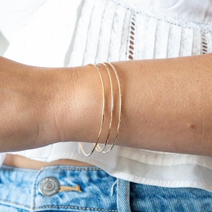 以下が含まれることがあります： 細いゴールドカラーのブレスレットが3つ、手首に重ねられています。細くて円形のブレスレットはシンプルでエレガントです。白いトップスと青いジーンズを着用しています。