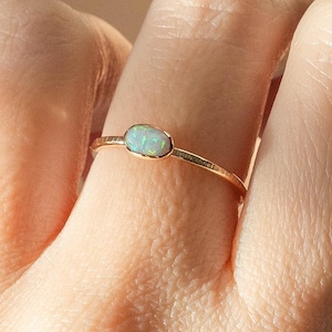 May include: A gold ring featuring an oval opal gemstone. The opal has a mix of green and blue hues. The ring's band has a textured design. The ring is shown on a finger.