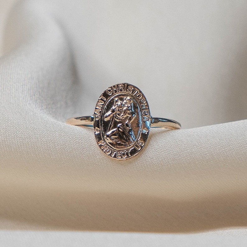Puede incluir: Un anillo dorado con un colgante ovalado que representa a San Crist&oacute;bal, con la inscripci&oacute;n "SAINT CHRISTOPHER PROTECT US". El anillo est&aacute; colocado sobre una tela de color crema.