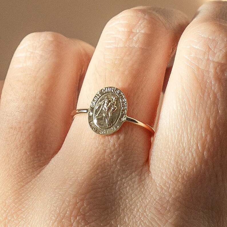 Puede incluir: Un anillo dorado con un colgante ovalado que representa a San Crist&oacute;bal. El anillo tiene una banda delgada y se lleva en un dedo. El colgante tiene la inscripci&oacute;n "SAINT CHRISTOPHER PROTECT US" en el borde.