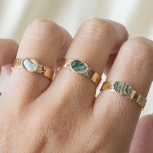 Pode incluir: Três anéis de ouro com bandas marteladas, cada um com uma incrustação oval de concha de abalone. As conchas iridescentes exibem tons de verde, azul e branco. Os anéis são exibidos nos dedos.