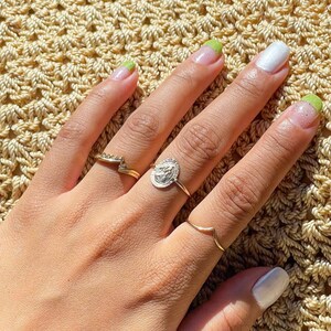 Puede incluir: Una mano con u&ntilde;as pintadas y varios anillos. Las u&ntilde;as est&aacute;n pintadas con una combinaci&oacute;n de colores blanco y verde. Los anillos son dorados y plateados, uno de ellos con un dise&ntilde;o de moneda.