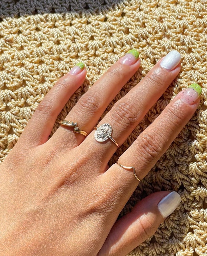 Puede incluir: Una mano con u&ntilde;as pintadas y varios anillos. Las u&ntilde;as est&aacute;n pintadas con una combinaci&oacute;n de colores blanco y verde. Los anillos son dorados y plateados, uno de ellos con un dise&ntilde;o de moneda.