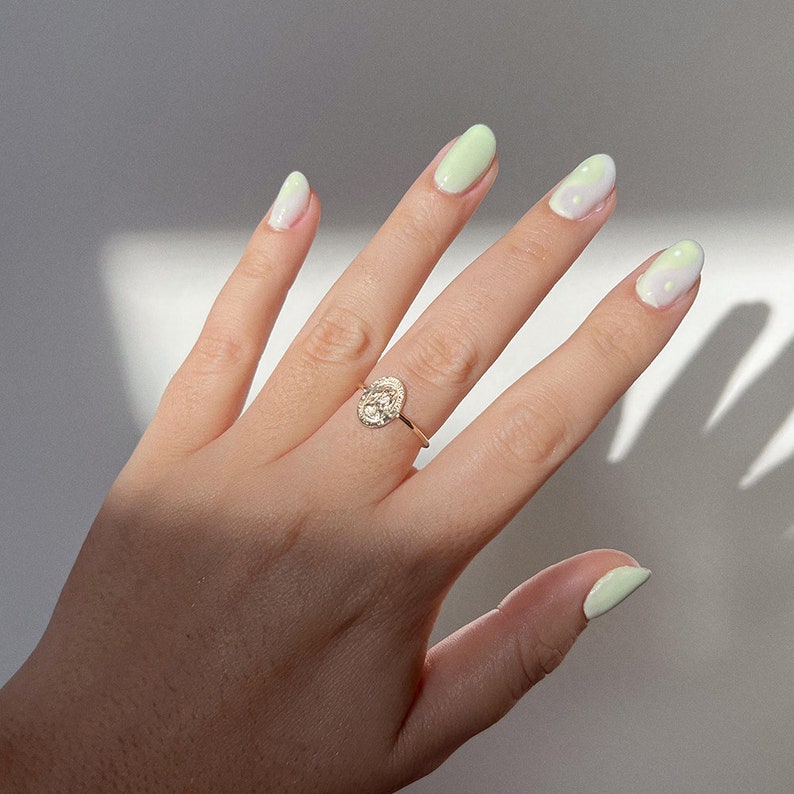 Puede incluir: Un anillo de oro con un diamante de forma ovalada rodeado de diamantes m&aacute;s peque&ntilde;os. El anillo est&aacute; en una mano con esmalte de u&ntilde;as degradado verde claro y blanco.