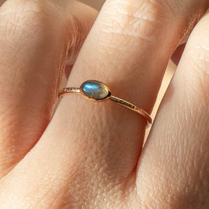 Puede incluir: Un anillo de oro con una pequeña piedra preciosa azul de forma ovalada. La banda del anillo tiene un diseño texturizado y martillado. La piedra preciosa está engastada en un bisel simple. El anillo se muestra en un dedo.