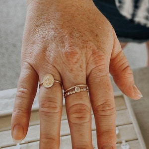 Puede incluir: Una mano con dos anillos de oro. Uno presenta un dise&ntilde;o de medall&oacute;n, y el otro tiene un estilo delicado y en capas con peque&ntilde;os adornos. La imagen incluye el texto "Love seeing our pieces on you guys!"