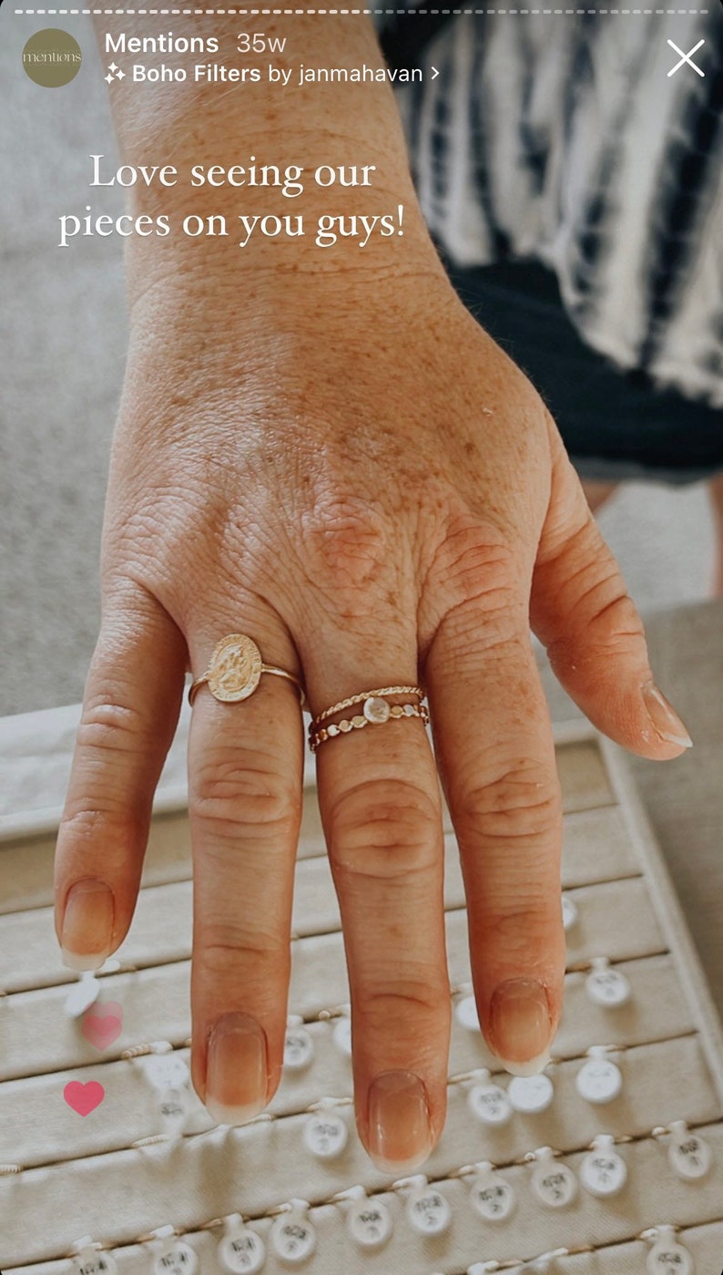 Puede incluir: Una mano con dos anillos de oro. Uno presenta un dise&ntilde;o de medall&oacute;n, y el otro tiene un estilo delicado y en capas con peque&ntilde;os adornos. La imagen incluye el texto "Love seeing our pieces on you guys!"
