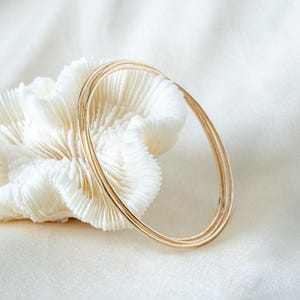 May include: Three delicate gold bangles, each with a thin, smooth band, are displayed against a backdrop of a white, textured coral-like object. The bangles are simple and elegant, reflecting light. The background is a soft, white fabric.
