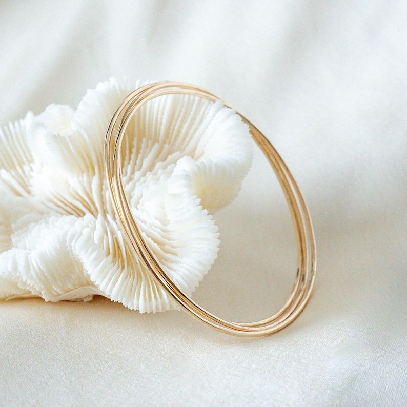 May include: Three delicate gold bangles, each with a thin, smooth band, are displayed against a backdrop of a white, textured coral-like object. The bangles are simple and elegant, reflecting light. The background is a soft, white fabric.