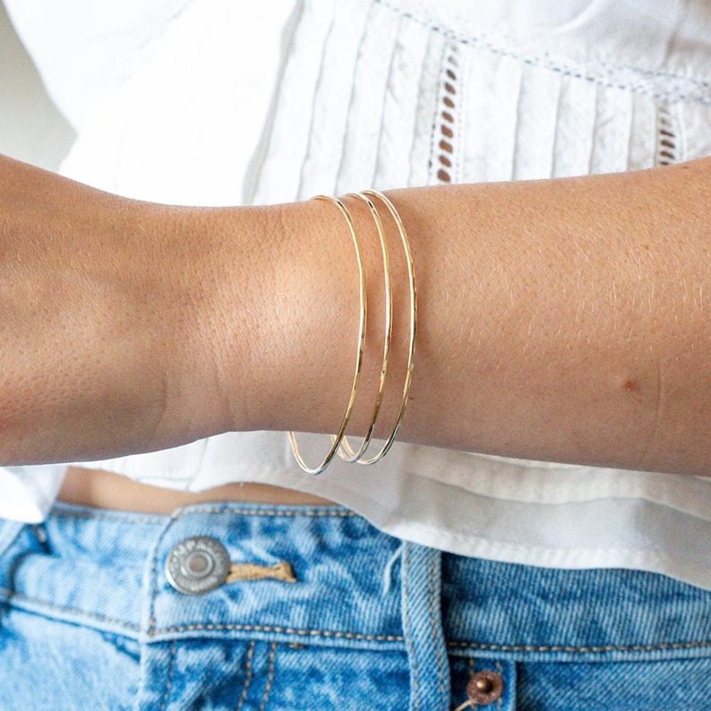 May include: Three delicate gold-colored bangles are stacked on a wrist. The thin, circular bracelets are simple and elegant. The person is wearing a white top and blue jeans.