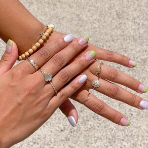 Puede incluir: Manos con u&ntilde;as manicuradas, con una combinaci&oacute;n de colores lila claro y verde. Las u&ntilde;as est&aacute;n adornadas con un dise&ntilde;o degradado. Tambi&eacute;n son visibles varios anillos de oro y una pulsera de cuentas de madera.