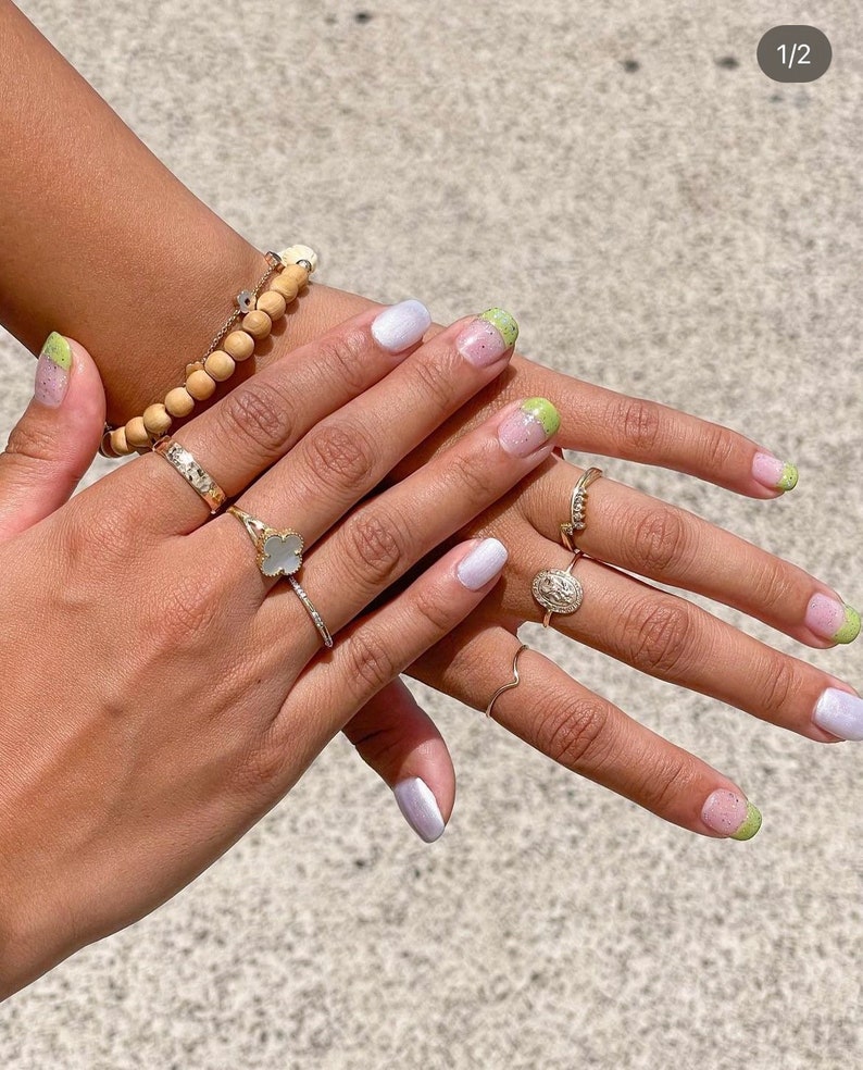 Puede incluir: Manos con u&ntilde;as manicuradas, con una combinaci&oacute;n de colores lila claro y verde. Las u&ntilde;as est&aacute;n adornadas con un dise&ntilde;o degradado. Tambi&eacute;n son visibles varios anillos de oro y una pulsera de cuentas de madera.