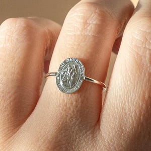 May include: A silver ring featuring an oval pendant with the image of Saint Christopher. The pendant is inscribed with the words "ST CHRISTOPHER PROTECT US". The ring is worn on a finger.