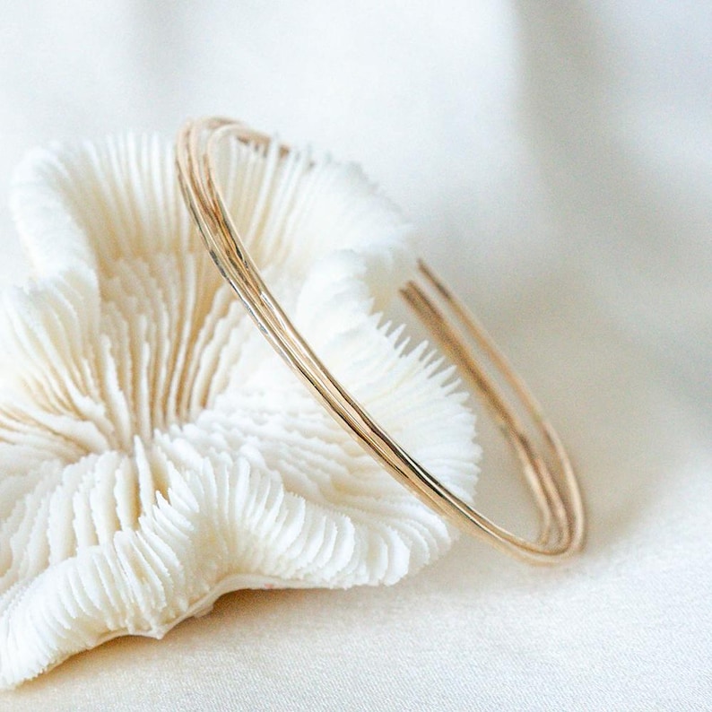 May include: Three delicate gold bangles rest on a white, textured coral-like object against a soft, white background. The bangles are thin and have a slightly hammered texture, reflecting light. The jewelry is simple and elegant.
