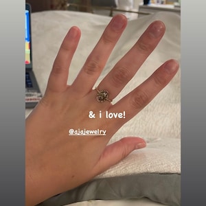 Puede incluir: Un anillo dorado con un dise&ntilde;o detallado se muestra en una mano. El anillo presenta una piedra central rodeada de peque&ntilde;os adornos. El texto en la imagen dice "& i love!" y "@ajajewelry".
