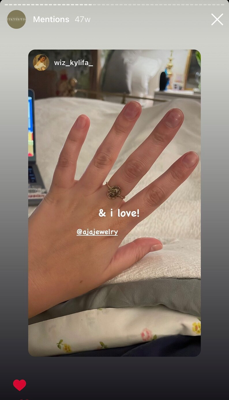 Puede incluir: Un anillo dorado con un dise&ntilde;o detallado se muestra en una mano. El anillo presenta una piedra central rodeada de peque&ntilde;os adornos. El texto en la imagen dice "& i love!" y "@ajajewelry".