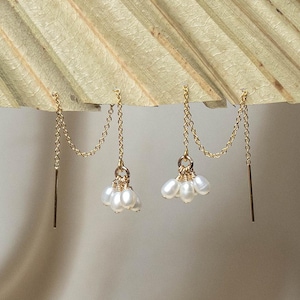 Puede incluir: Un par de pendientes colgantes dorados. Cada pendiente presenta una fina cadena conectada a un grupo de perlas blancas y una barra recta. Los pendientes se muestran sobre un fondo neutro, resaltando su elegante diseño.