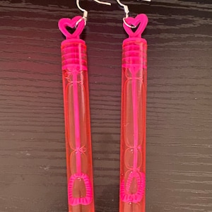 May include: A pair of pink bubble wands with heart-shaped tops and silver hooks. The wands are made of clear plastic and have a pink liquid inside.