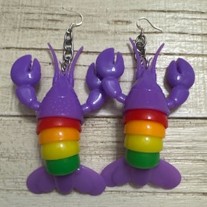 May include: A pair of purple lobster earrings with a rainbow stripe design. The earrings are made of plastic and have silver hooks.