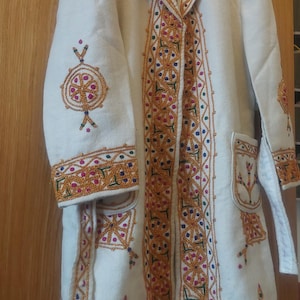 Op de afbeelding: Een witte jas met ingewikkelde borduursels in goud, blauw, groen en roze. De jas heeft een kraag en een knoopsluiting. De borduursels bevinden zich op de kraag, manchetten en de voorkant van de jas.