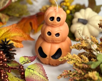 Puede incluir: Una vela de calabaza apilada con tres calabazas naranjas, cada una con una cara de calabaza tallada. La vela está rodeada de follaje de otoño.