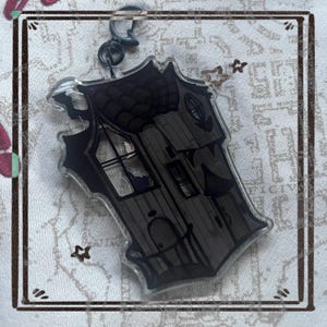 May include: A clear acrylic keychain with a black and grey illustration of a haunted house. The house has a detailed roof, windows, and a door. The keychain is attached to a silver-coloured clasp. The overall design has a spooky theme.