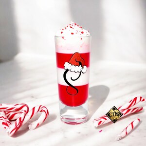 Puede incluir: Un vaso de chupito a capas con rayas rojas y blancas, cubierto con crema batida y chispas rojas. Un gráfico de gorro de Papá Noel está en el vaso. Bastones de caramelo están esparcidos alrededor del vaso.
