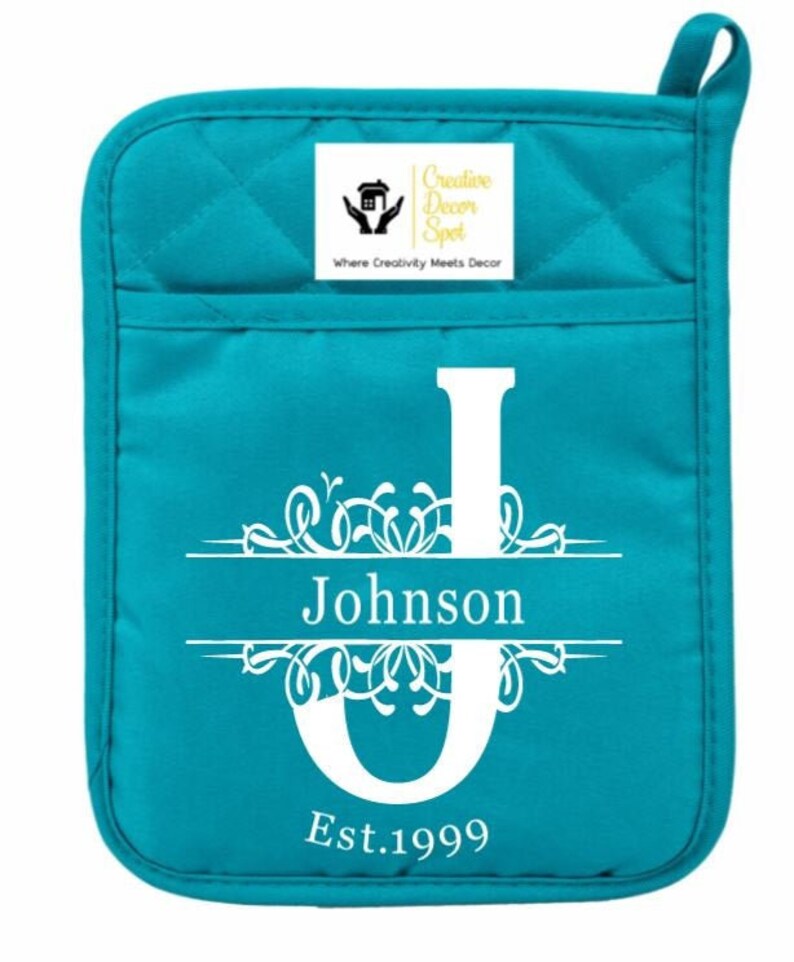 May include: Teal blue quilted oven mitt with a white monogram design. The monogram features a large letter "J" with a decorative flourish and the name "Johnson" centered below the letter. The text "Est. 1999" is centered below the name. A small white square label with the text "Creative Decor Spot" and the tagline "Where Creativity Meets Decor" is in the upper right corner.