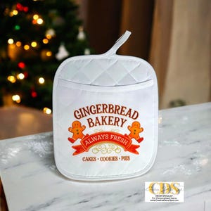 May include: White quilted oven mitt with a pocket and hanging loop. The mitt features the text "Gingerbread Bakery Always Fresh" with gingerbread man graphics and the words "Cakes, Cookies, Pies."