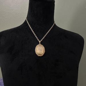 May include: A gold-colored oval locket necklace with a detailed filigree design. The locket hangs from a delicate chain, displayed on a black mannequin. The necklace is a vintage-inspired piece.