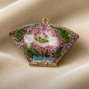 May include: A fan-shaped metal pendant featuring cloisonné enamel work with a central pink and white flower, green leaves, and a purple speckled background. The piece has a gold-toned metal frame, a small top loop for hanging, and a bottom hinge.