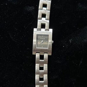 May include: Silver-toned Folio wristwatch with a square watch face and a linked band. The watch face is dark with silver hands and the brand name "Folio" printed at the top. The band is made of rectangular links.