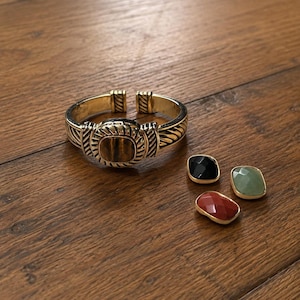 Op de afbeelding: Een goudkleurige manchetarmband met een centrale tijgeroogsteen en een gedraaide kabelstructuur. Drie extra verwisselbare rechthoekige stenen in zwart, lichtgroen en roodbruin zijn naast de armband geplaatst.
