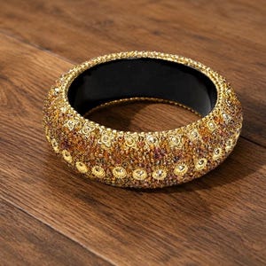 May include: A gold-colored bangle bracelet with a black interior. The bracelet is heavily embellished with small, sparkling stones in shades of gold, amber, and red. The bracelet is resting on a wooden surface.