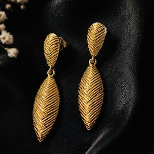 May include: A pair of gold-toned drop earrings featuring a textured, leaf-shaped design. Each earring consists of a smaller, teardrop-shaped top piece connected to a larger, elongated, pointed oval pendant, both displaying a detailed herringbone pattern.