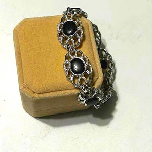 May include: Silver-toned bracelet with oval black stones set in ornate, openwork links. The bracelet is displayed on a small, square, tan jewelry box, creating a contrast of textures and colors. A classic piece of jewelry.