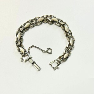 May include: A silver-toned metal bracelet featuring a double-chain design with alternating smooth and textured diamond-shaped links. The bracelet includes a box clasp closure and a small safety chain with a spring ring clasp for secure fastening.
