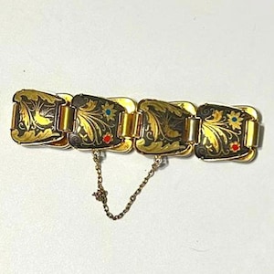 May include: Gold-toned bracelet with rectangular links. Each link features a black background with gold floral and leaf designs, accented with red and blue details. A delicate chain secures the bracelet.