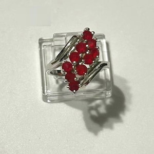 Peut inclure: Une bague en argent ornée d'un groupe de pierres précieuses rouges vives. Les pierres sont disposées en forme fluide, créant un design attrayant. La bague est présentée sur un support carré transparent sur fond blanc.