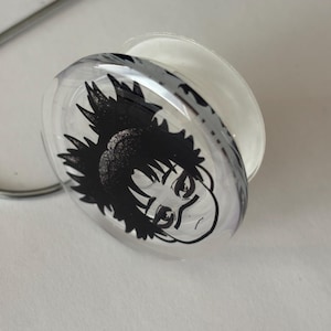 May include: A circular phone grip featuring a clear, glossy top layer with a black and white graphic illustration of an anime-style character with spiky hair and a headband. The base of the phone grip is white and collapsible in design.
