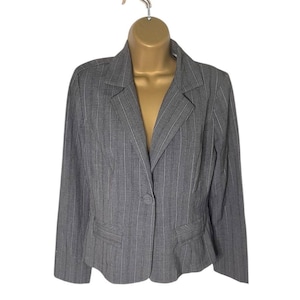 Puede incluir: Un blazer gris con finas rayas blancas, exhibido en un maniquí. El blazer tiene solapas con muescas, dos bolsillos delanteros y un cierre de un solo botón. Las mangas largas y el corte entallado sugieren un estilo profesional o de negocios.