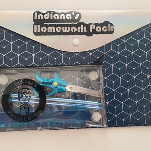 May include: A blue and white geometric patterned folder with a clear plastic pouch attached. The pouch contains a pair of scissors, a ruler, and pencils. The folder has a silver iridescent flap with the text "Indiana's Homework Pack" printed on it.
