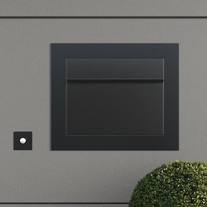 May include: A black mailbox with a white button doorbell mounted on a gray wall. A green bush is in the foreground.