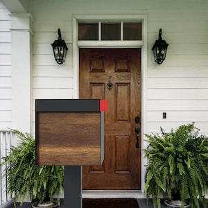May include: A black mailbox with a woodgrain finish and a red accent on the top. The mailbox is mounted on a black metal post and is in front of a white house with a wooden door.